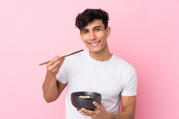 Jovem argentino sobre parede branca isolada, segurando uma tigela de macarrão com pauzinhos