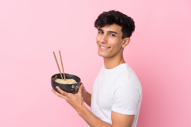 Jovem argentino sobre parede branca isolada, segurando uma tigela de macarrão com pauzinhos