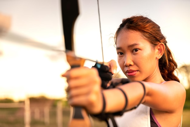 Jovem arqueira feminina atirar em flecha com arco no campo da natureza para atingir o conceito de sucesso no campo para exercício esportivo na hora do pôr do sol