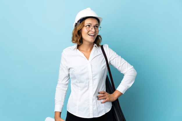 Jovem arquiteta georgiana com capacete segurando plantas na parede isolada, posando com os braços na cintura e sorrindo