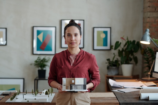 Foto jovem arquiteta ou designer contemporânea em trajes casuais segurando um modelo de papel da nova casa enquanto está em um ambiente de escritório