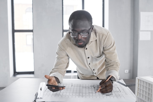 Foto jovem arquiteto africano usando seu telefone celular enquanto desenha um projeto na mesa do escritório