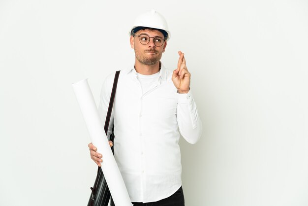 Jovem arquiteto com capacete e segurando plantas isoladas no fundo branco com dedos cruzando e desejando o melhor