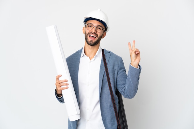 Jovem arquiteto com capacete e segurando plantas isoladas no fundo branco sorrindo e mostrando o sinal da vitória