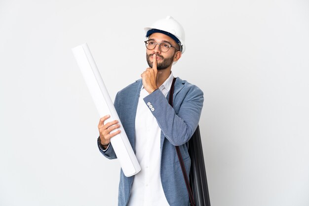 Jovem arquiteto com capacete e segurando plantas isoladas no fundo branco, tendo dúvidas enquanto olha para cima