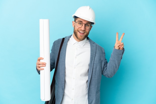 Jovem arquiteto com capacete e segurando plantas isoladas sorrindo e mostrando sinal de vitória