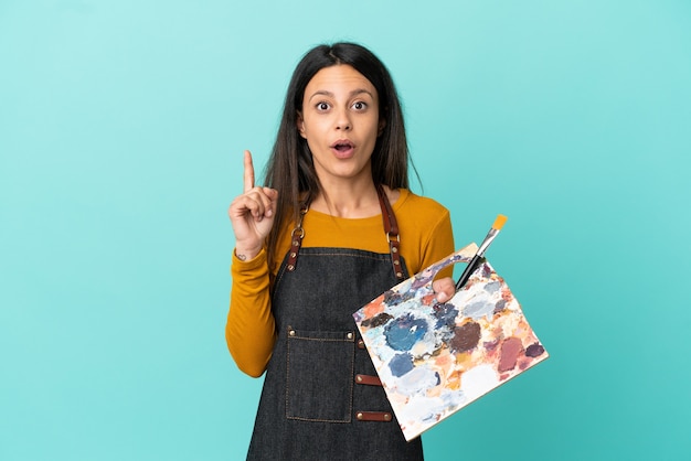 Jovem artista caucasiana segurando uma paleta isolada em um fundo azul com a intenção de perceber a solução enquanto levanta um dedo