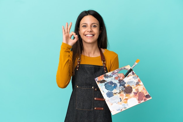 Jovem artista caucasiana segurando uma paleta isolada em um fundo azul e mostrando um sinal de ok com os dedos