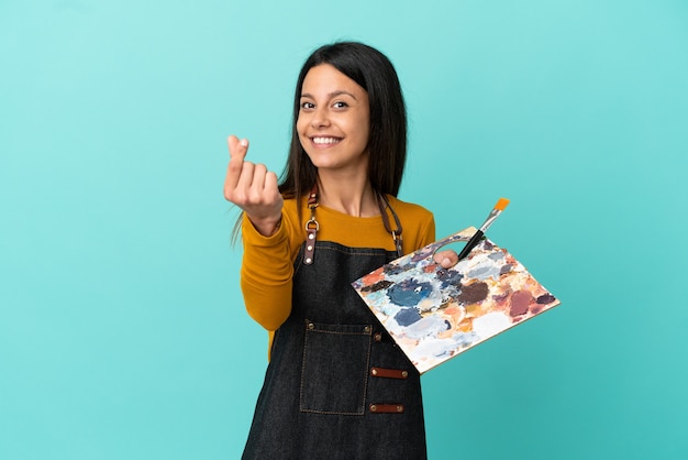 Jovem artista caucasiana segurando uma paleta isolada em um fundo azul fazendo gesto de dinheiro