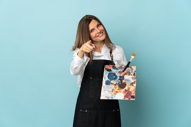 Jovem artista eslovaca isolada em um fundo azul apontando para a frente com uma expressão feliz