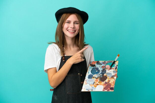 Jovem artista inglesa segurando uma paleta isolada em fundo azul apontando para o lado para apresentar um produto