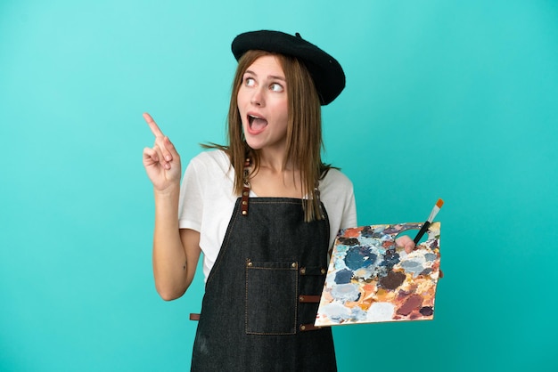 Foto jovem artista inglesa segurando uma paleta isolada em fundo azul com a intenção de perceber a solução enquanto levanta um dedo