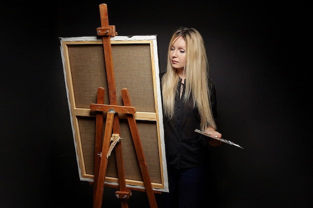 Jovem artista loira está atrás de um cavalete. sessão de fotos em um fundo preto no estúdio.