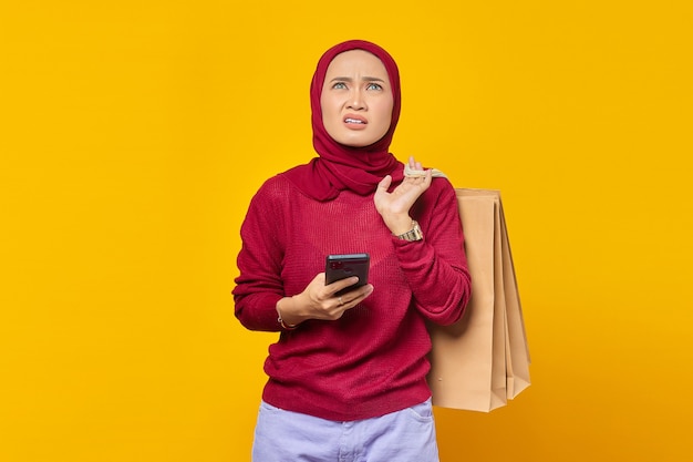 Jovem asiática chocada segurando uma sacola de compras e um smartphone enquanto olha para cima sobre um fundo amarelo