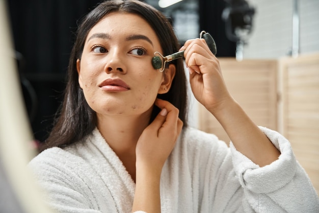 Foto jovem asiática com cabelo castanho usando rolo de jade para uma massagem facial na casa de banho auto-cuidados