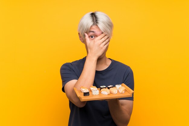 Jovem asiática com sushi sobre amarelo isolado cobrindo os olhos e olhando por entre os dedos