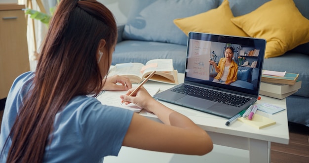 Jovem asiática com videochamada para computador portátil de uso casual, aprender on-line com o professor, escrever palestra, caderno, sala de estar em casa