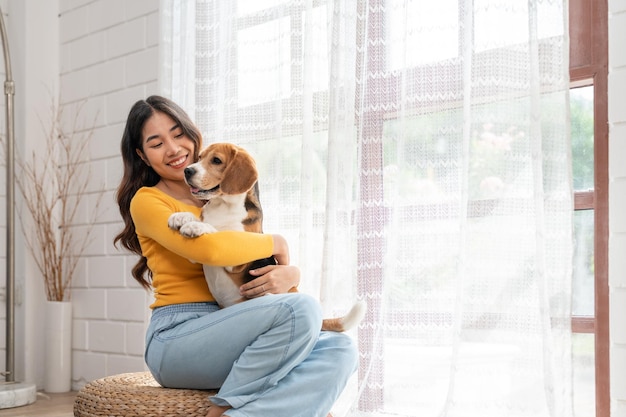 Jovem asiática em roupas casuais abraçando seu cachorro beagle sentado na sala de estar em casa