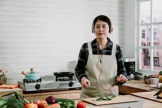 Jovem asiática está de pé na cozinha preparando salada com frutas e legumes frescos. esposa em fones de ouvido brancos ouve música. mulher cortando pepino em fatias na tábua de madeira.