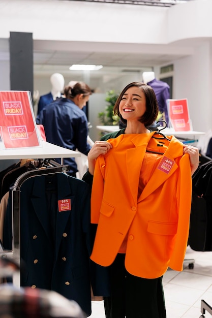 Foto jovem asiática feliz e excitada segurando um item de roupa no gancho nas mãos e sorrindo felizmente. cliente feminina se sentindo satisfeita com os preços baixos enquanto faz compras na sexta-feira negra no centro comercial de moda