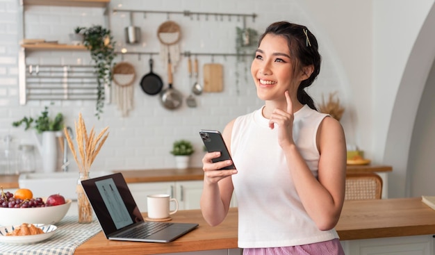 Jovem asiática feliz e interessada em pensar usando telefone celular na cozinha