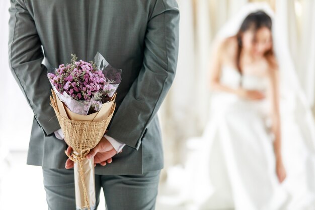 Jovem asiática linda feliz cabelo comprido noiva vestido de noiva branco com véu de renda seethrough sentar sorriso esperando o noivo de terno cinza que esconde o buquê de flores nas costas para surpreender no camarim.