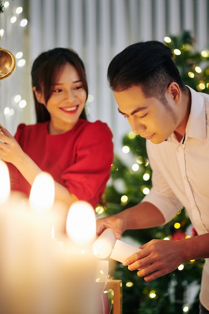 Jovem asiática muito sorridente olhando para o namorado acendendo velas ao decorar um apartamento para o Natal