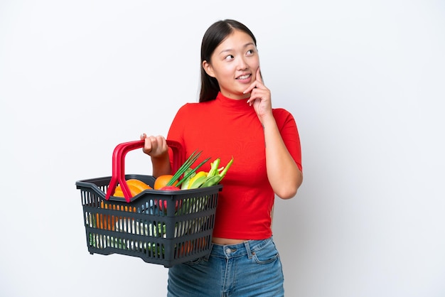 Jovem asiática segurando uma cesta de compras cheia de comida isolada em fundo branco pensando em uma ideia enquanto olha para cima