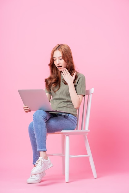 Jovem asiática sentada e usando laptop em fundo rosa