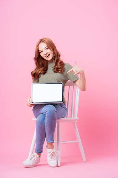 Jovem asiática sentada e usando laptop em fundo rosa