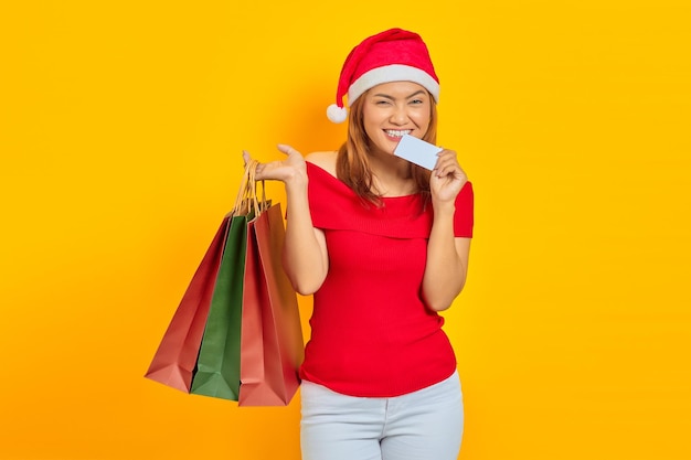 Jovem asiática sorridente com chapéu de Papai Noel segurando uma sacola de compras e mordendo o cartão de crédito