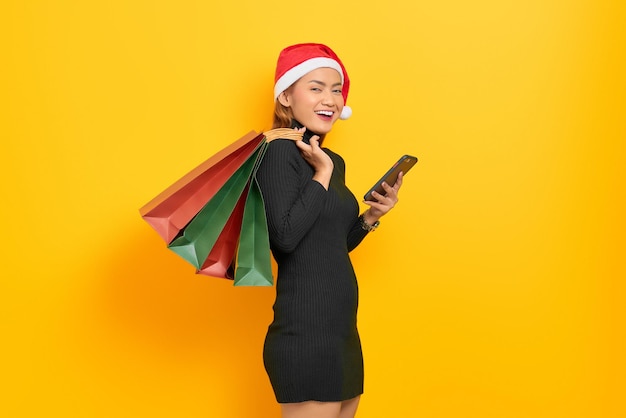 Jovem asiática sorridente com chapéu de Papai Noel, usando um telefone celular e segurando sacolas de compras isoladas sobre fundo amarelo