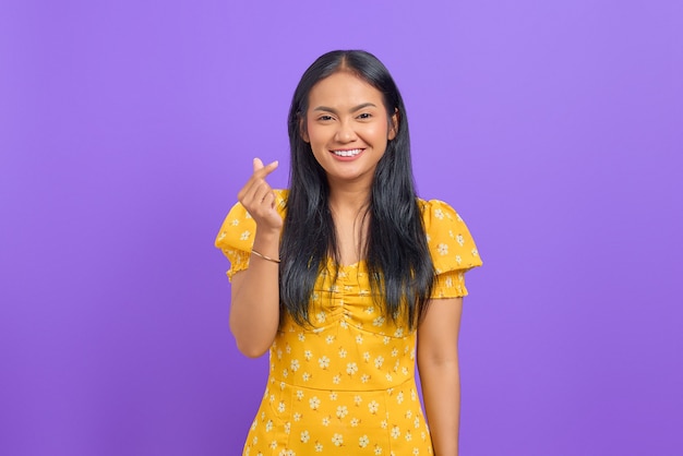 Jovem asiática sorridente mostrando o sinal do coração com os dedos cruzados no fundo roxo