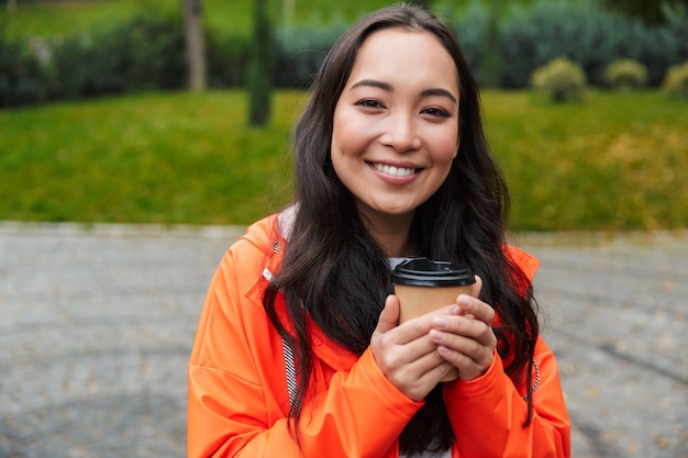 Jovem asiática sorridente, vestindo uma capa de chuva, caminhando ao ar livre na chuva, segurando uma xícara de café