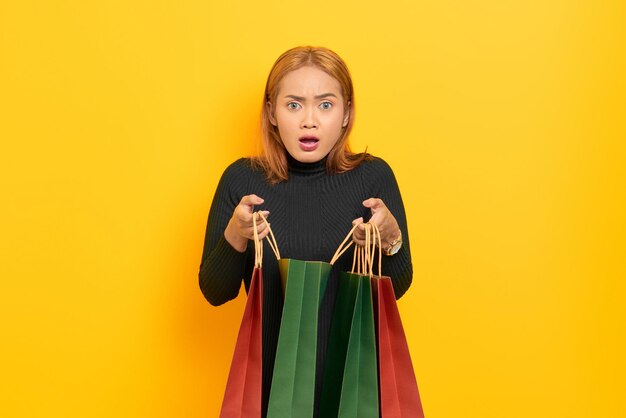 Jovem asiática surpreendida abrindo sacolas de compras com a boca aberta isolada sobre fundo amarelo