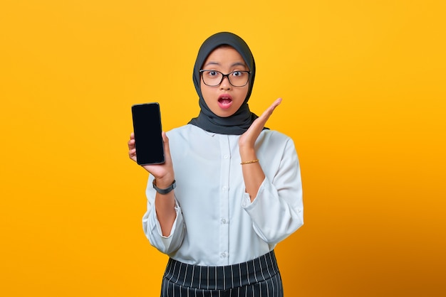 Jovem asiática surpreendida mostrando a tela em branco do celular isolada sobre um fundo amarelo