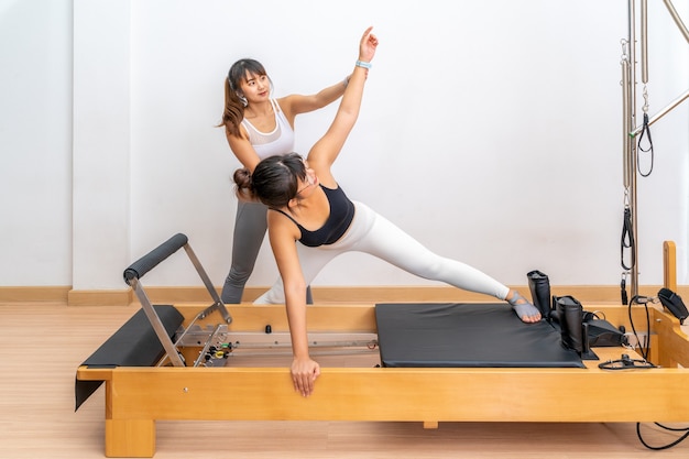 Jovem asiática trabalhando em uma máquina reformadora de pilates com sua treinadora durante o treinamento de exercícios de saúde