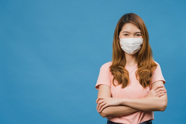 Jovem asiática usando máscara médica com os braços cruzados, vestida com uma roupa casual e olhando para a frente, isolada na parede azul