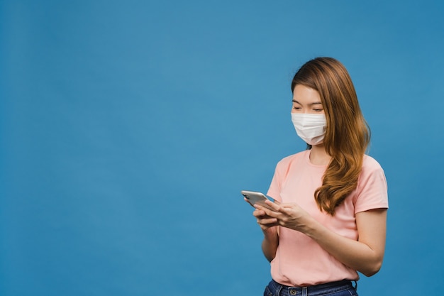 Jovem asiática usando máscara médica usando um telefone celular com roupas casuais isoladas na parede azul