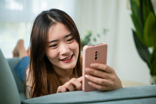 Jovem asiática usando telefone celular inteligente deitado no sofá