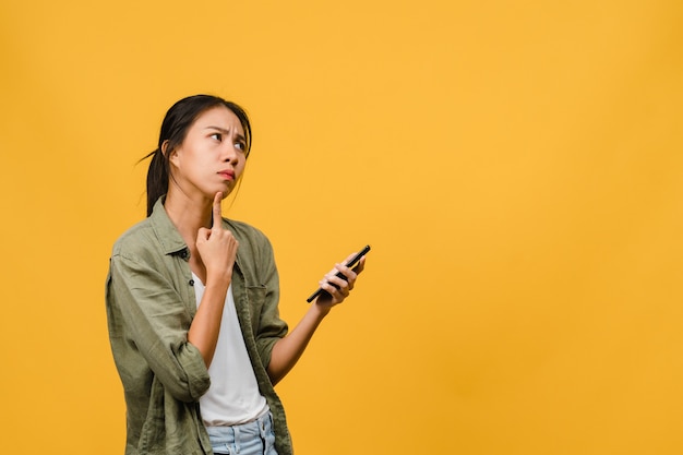 Jovem asiática usando telefone com expressão positiva, vestida com um pano casual na parede amarela