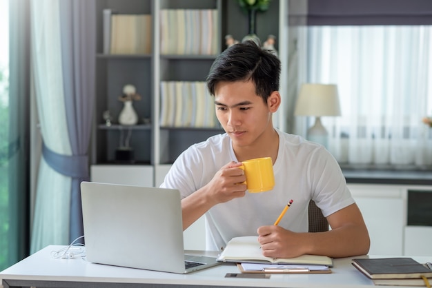 Jovem asiático bebendo café e fazer anotações, trabalhar em casa, olhando para o laptop.