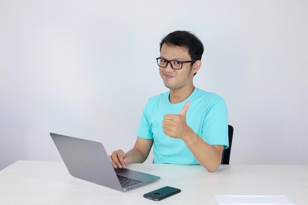 Jovem asiático bonito aparecendo polegar enquanto trabalhava com laptop indonésio vestindo camisa azul