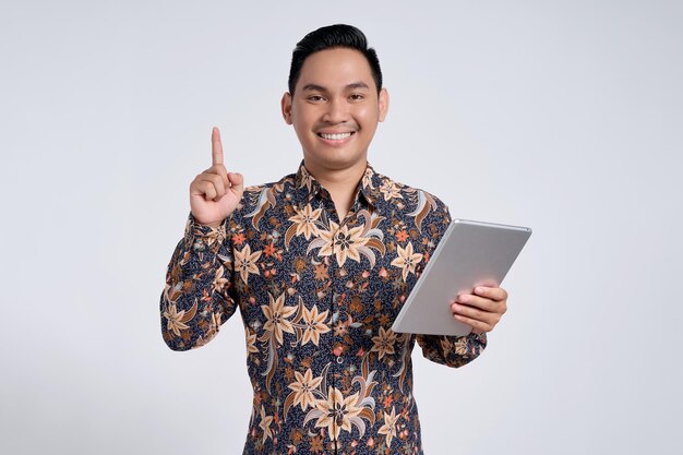 Jovem asiático bonito e excitado vestindo camisa batik segurando tablet digital e apontando o dedo para cima recebendo ideias depois de ler um artigo isolado em fundo branco conceito de estilo de vida das pessoas