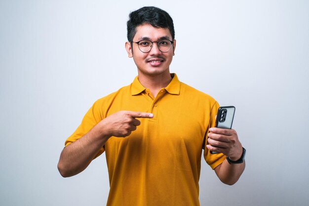 Jovem asiático bonito usando smartphone vestindo camisa casual isolado fundo branco muito feliz apontando com a mão e o dedo