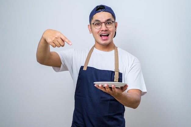 Jovem asiático bonito vestindo avental mostrando expressão animada enquanto segura o prato vazio