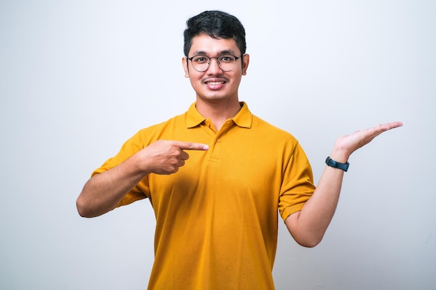 Jovem asiático bonito vestindo camisa casual sobre fundo branco espantado e sorrindo para a câmera ao apresentar com a mão e apontando com o dedo