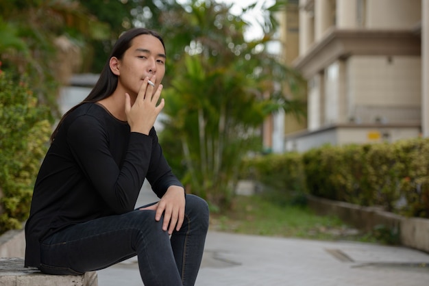 Jovem asiático com cabelo comprido sentado e fumando no parque