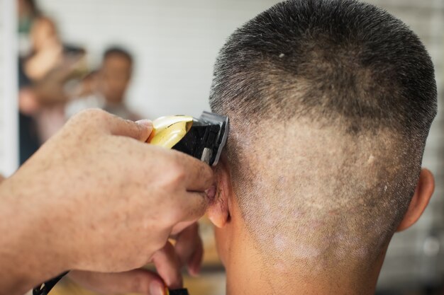 Jovem asiático com cabelos grisalhos, sendo cortado com uma máquina de tosquia elétrica por um barbeiro profissional na barbearia.