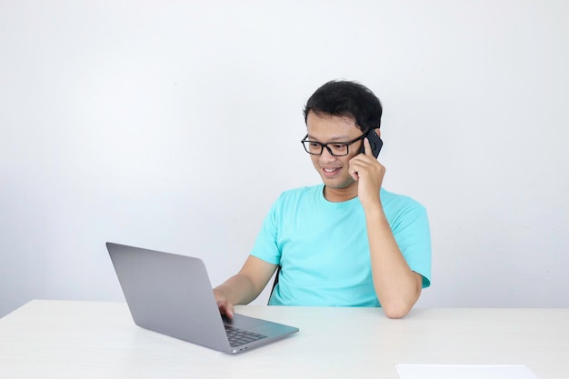 Jovem asiático com cara feliz está falando em um telefone celular com laptop na mesa homem indonésio vestindo camisa azul